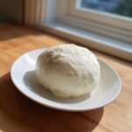 A smooth, white ball of fufu resting on a small white plate, illuminated by natural light.