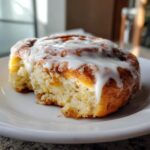 A close-up of a soft, fluffy cinnamon roll with thick white icing dripping down the side, sitting on a white plate.