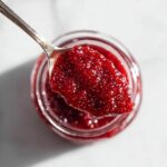Overhead view of a spoonful of thick, vibrant red raspberry jam being lifted from a glass jar.