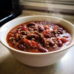 Close-up of a steaming white bowl filled with rich, hearty crockpot chili featuring ground beef and kidney beans.
