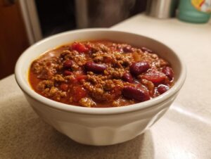 A close-up shot of a steaming white bowl filled with rich, hearty crockpot chili featuring ground beef and kidney beans.
