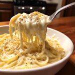 A fork lifting spaghetti heavily coated in rich, creamy homemade alfredo sauce, showing a cheese pull and black pepper.