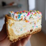 A hand holding a thick slice of sugar cookie cheesecake filled with colorful sprinkles and topped with whipped cream and more sprinkles.