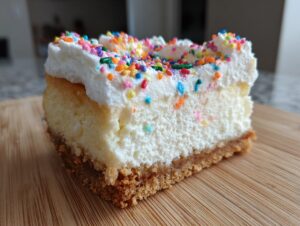 A close-up of a slice of sugar cookie cheesecake topped with whipped cream and colorful sprinkles.