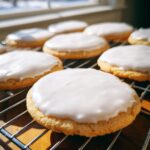 Close-up of round sugar cookies topped with smooth, white sugar cookie icing, cooling on a wire rack near a window.