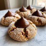 Several freshly baked peanut butter kiss cookies, rolled in sugar and topped with a chocolate kiss.