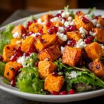 Close-up of a vibrant sweet potato salad featuring roasted orange sweet potato cubes, crumbled white feta cheese, and bright red pomegranate seeds over greens.