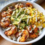 A vibrant shrimp bowl featuring grilled shrimp, diced avocado, corn, and a creamy white sauce drizzled over white rice.