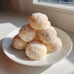 A stack of powdered sugar-dusted lemon cookies on a white plate, backlit by sunlight near a window.