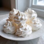 A close-up of several perfectly piped, bright white meringue cookies stacked on a white plate near a window.