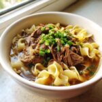 Close-up of a white bowl filled with rich broth, wide noodles, shredded beef, and topped with fresh green onions for amazing beef noodle soup.