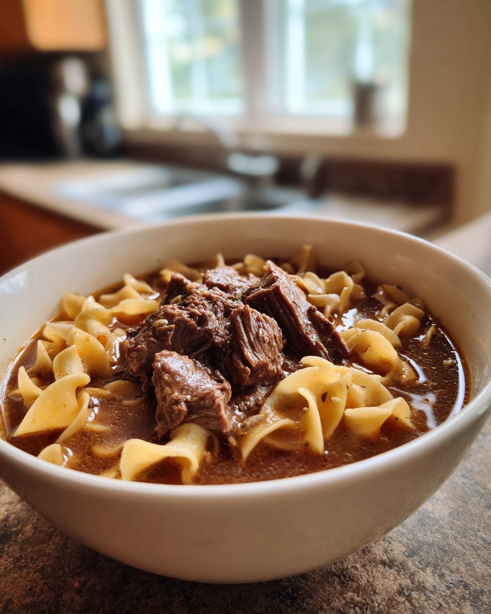 Close-up of a white bowl filled with rich beef noodle soup, featuring tender chunks of beef and egg noodles.