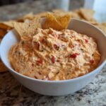 Close-up of creamy, orange-tinted boat dip mixed with red peppers, served in a white bowl with tortilla chips.