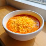 A white bowl filled with hot, creamy chili cheese dip, generously topped with shredded cheddar cheese.