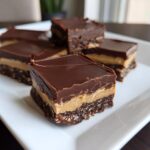 Five decadent peanut butter bars showing distinct layers of chocolate topping, creamy peanut butter, and a crumbly base, served on a white plate.