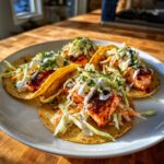 Four fully loaded salmon tacos with seasoned fish, slaw, and white sauce on a white plate.