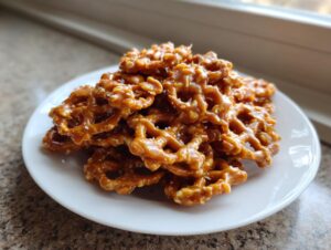 A close-up of a pile of crunchy toffee pretzels coated in glossy caramel and sprinkled with sea salt on a white plate.
