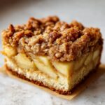 A close-up, tempting shot of a square slice of apple crumb cake showing layers of soft cake, thick apple filling, and a crunchy cinnamon topping.