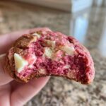 Close-up of a hand holding a bitten raspberry cheesecake cookies, revealing a vibrant pink, soft interior with white chocolate chips.