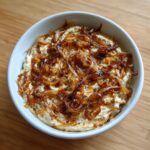 A white bowl filled with creamy caramelized onion dip, generously topped with dark, crispy caramelized onions.
