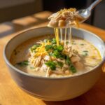 A spoonful of creamy chicken chili, topped with shredded cheese and cilantro, shows melted cheese strings dripping back into the bowl of chicken chili.