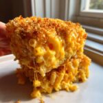 A hand lifts a square portion of baked mac and cheese showing the gooey interior and crispy topping.
