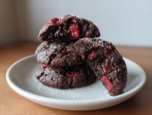 A stack of rich, dark chocolate cherry cookies, one broken open to show gooey chocolate and bright red cherries.