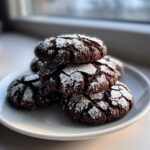 A stack of five rich, dark chocolate crinkle cookies dusted heavily with white powdered sugar on a white plate.