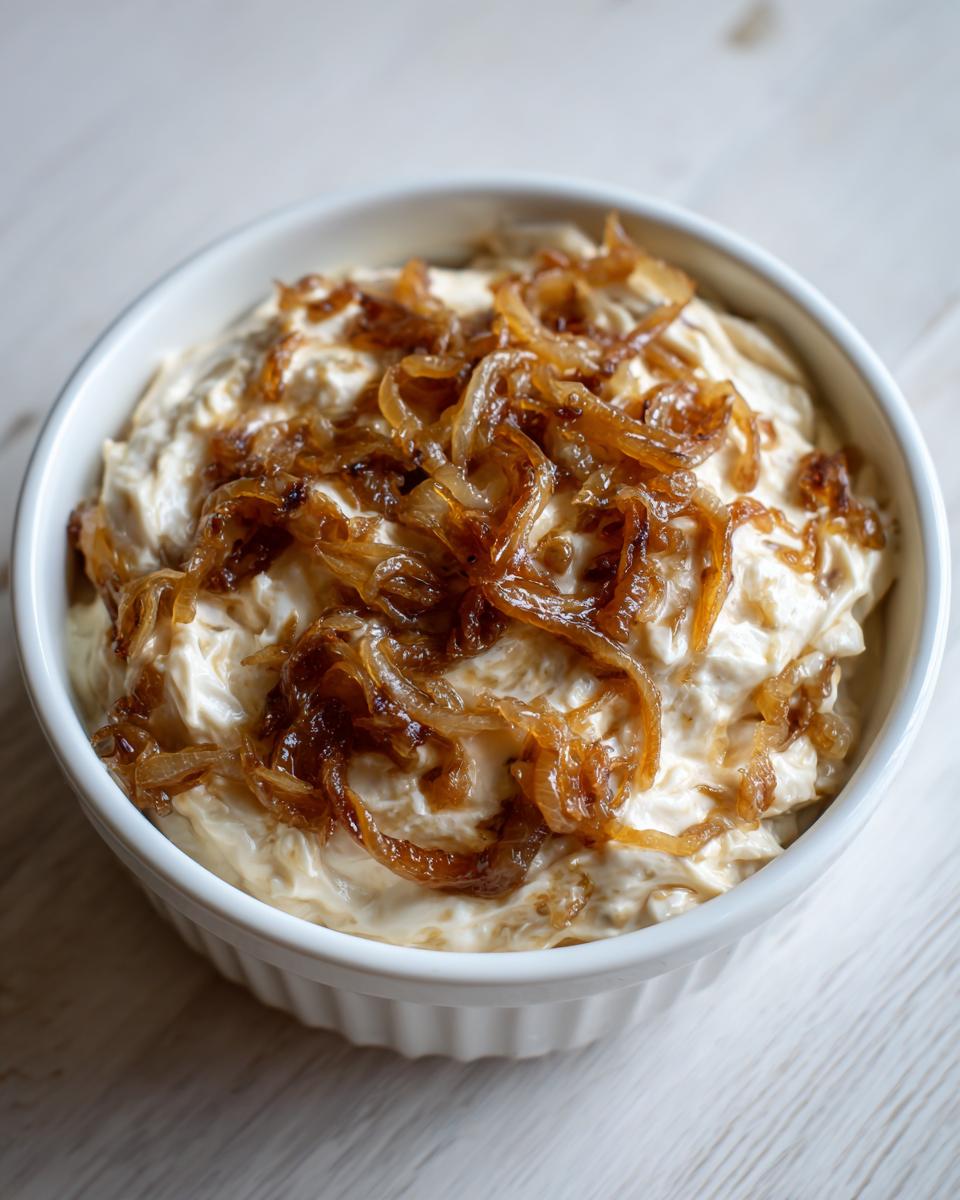 Close-up of a white bowl filled with creamy caramelized onion dip, generously topped with golden brown caramelized onions.