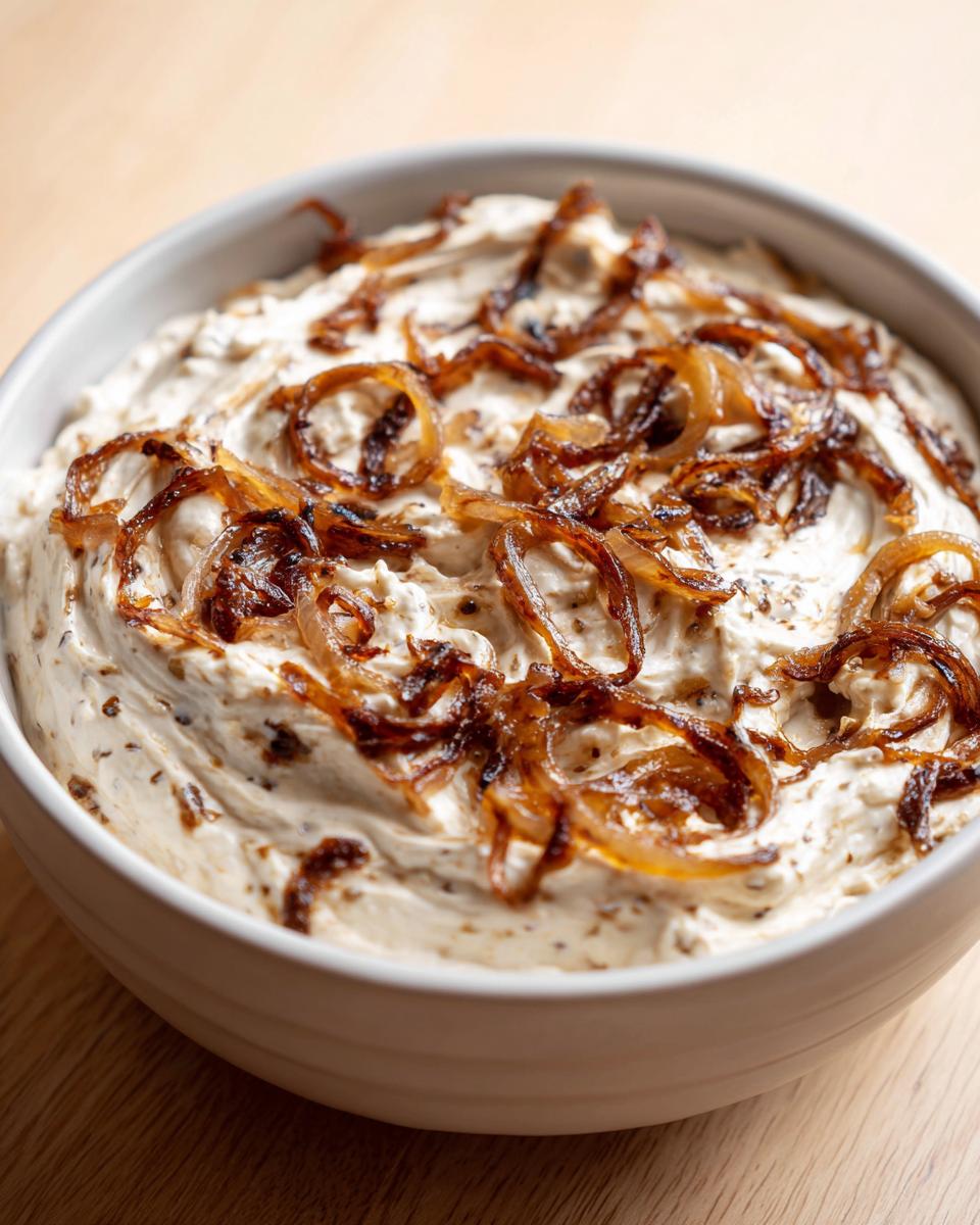 A close-up of creamy caramelized onion dip topped generously with crispy, dark caramelized onion rings.