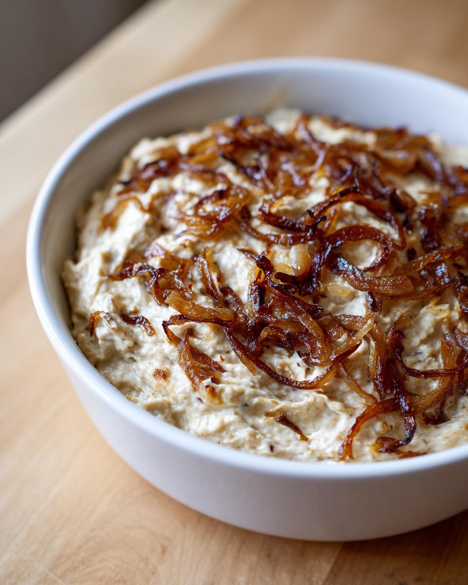 Close-up of a white bowl filled with creamy caramelized onion dip, generously topped with dark, crispy caramelized onions.