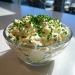 A close-up of creamy, white clam dip generously topped with freshly chopped green chives in a clear glass bowl.