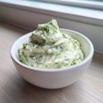 A close-up of creamy, pale green dill dip mounded high in a white bowl, garnished with fresh dill.