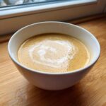 A close-up of a bowl of creamy roasted garlic soup, swirled with cream, sitting on a wooden surface.