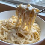 A fork lifting a generous portion of pasta coated in rich, creamy olive garden alfredo sauce from a white bowl.
