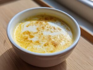 A close-up of creamy, yellow roasted garlic soup topped with cream swirl and spice in a white bowl.