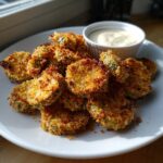 A pile of golden brown, crispy Fried Pickles served on a white plate next to a small ramekin of creamy dipping sauce.
