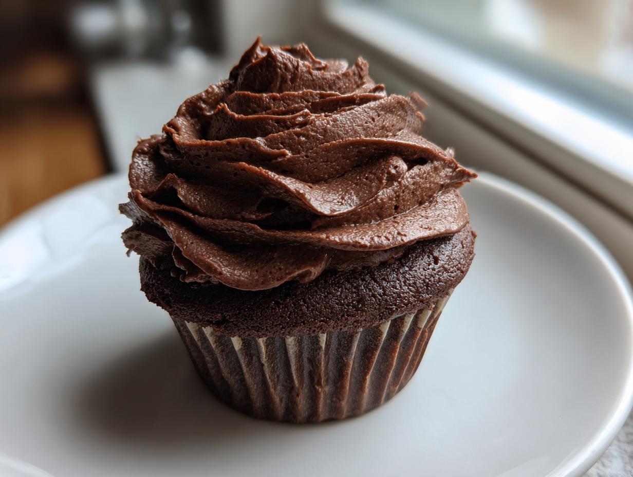 A close-up of a dark chocolate cupcake topped with a generous swirl of rich chocolate frosting.