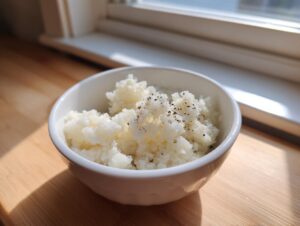 A close-up of fluffy cauliflower rice seasoned with black pepper, served in a white bowl on a wooden surface.