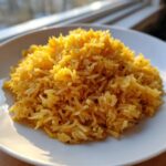A close-up of fluffy, golden yellow turmeric rice piled high on a white plate, brightly lit by sunlight.