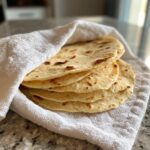 A stack of warm, freshly cooked corn tortillas wrapped partially in a white cloth on a granite countertop.