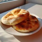 Two golden-brown, freshly baked rounds of pita bread stacked slightly on a white plate, illuminated by sunlight.