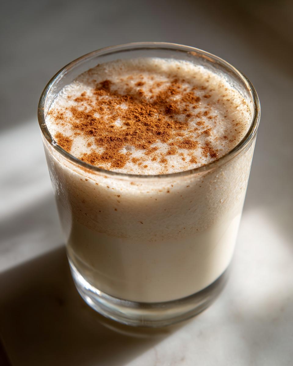 Close-up of a glass filled with creamy coquito, garnished generously with ground cinnamon on top.