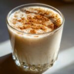 Close-up of a glass filled with creamy coquito, garnished generously with ground cinnamon on top.