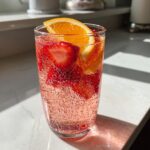 Close-up of a tall glass filled with fizzy pink champagne punch, strawberries, and an orange slice garnish.