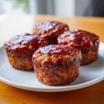 Four perfectly baked mini meatloaf portions topped with a shiny, thick red glaze, served on a white plate.