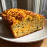 A golden-brown loaf of moist mexican cornbread, studded with melted cheddar cheese and green jalapeño pieces, resting on a white plate.