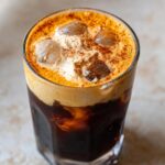 Close-up of a glass filled with pumpkin cream cold brew, featuring dark coffee, ice cubes, and a thick layer of orange-tinted cream topped with cinnamon.