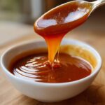 Close-up of thick, glossy caramel sauce dripping from a spoon back into a white bowl.