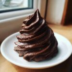 A tall, perfectly piped swirl of rich, dark chocolate frosting on a small white plate.